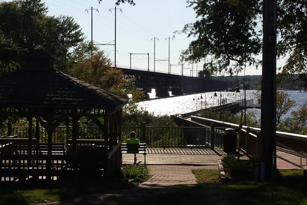 View from behind park on shore with long walkway extending out toward water. Bridge on left of water
